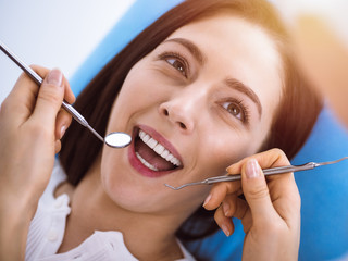 Smiling brunette woman being examined by dentist at sunny dental clinic. Hands of a doctor holding dental instruments near patient's mouth. Healthy teeth and medicine concept