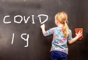 Little girl writing COVID on black chalkboard
