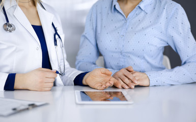 Unknown woman-doctor is showing to her patient a description of medication, while sitting together at the desk in the cabinet in a clinic. Female physician is using a computer tablet and a stethoscope