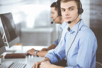 Two young men in headset, sitting at the desk in the modern office, listening to the clients. Call center operators at work