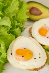 Two fried eggs for healthy breakfast on white plate with fresh avocado and green salad. Prepared toast with vegetables and tasty eggs  on white wooden background. 