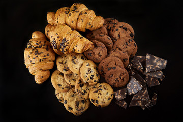 Tasty golden croissants on black background. Freshly baked chocolate chip cookies with broken milk chocolate pieces. Closeup of a group of French pastry from bakery .Top view with Copy space. 