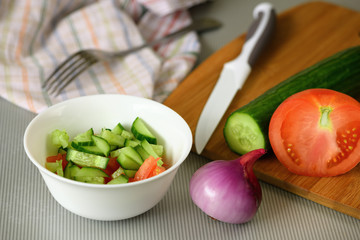 One white glass bowl with the pieces of the fresh food, the vegetables, one knife and one wooden cutting board are lying on the gray kitchen table.