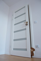 A brown teddy bear smiling is standing and looking into the room. Open white door and lavender on the door handle.