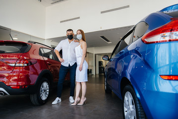 Young couple in masks selects a new vehicle and consult with a representative of the dealership in...