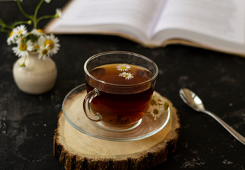 English tea with chamomile stands on a wooden frame and a teaspoon and an open book for reading lies on a black background. Nearby is a white vase with summer chamomile flowers