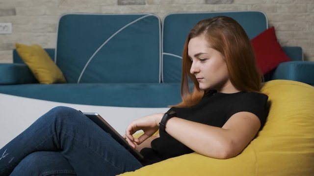 Young woman is sitting on a yellow bean bag and working at the tablet. Work from home concept.