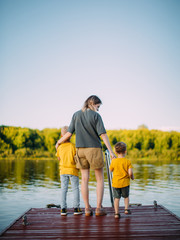 Cool baby boys with mother stand on pier by river. Back view. Summer photography for blog or ad about family and travel