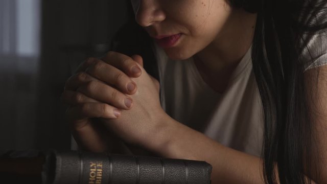 A woman prays with her hands crossed over a Bible for God's blessing in a dark room.