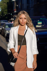 A european young beautiful blonde businesswoman in a white jacket is standing against the background of a road with cars. Dressed in business style