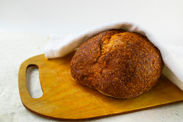 Freshly baked cereal bread on a wooden board under a white towel.