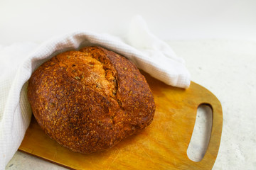 Freshly baked cereal bread on a wooden board under a white towel.