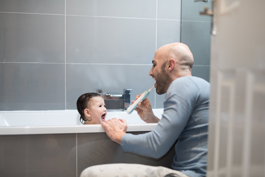 A Spanish man wearing blue pajamas brushes his teeth with an electric toothbrush as her daughter has a bath and laughs in bathroom in a bathroom with grey tiles in a house in Edinburgh, Scotland, Unit