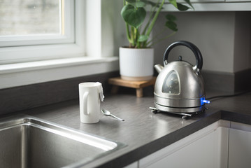 A boiling kettle next to a white cup with a tea bag on it and a spoon on a kitchen top next to a window in  a house Edinburgh, Scotland, United Kingdom, with a plant on the background