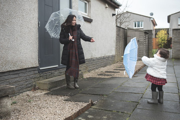 An Asian mother wearing a long jacket, Wellington boots and a long scarf holds a white umbrella and interacts with her daughter carrying a small blue umbrella as they smile outside a house in Edinburg