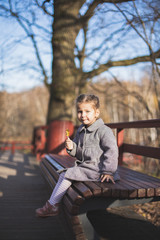 Elegant stylish little girl with pigtails in trendy grey coat, tights, snakers eats lollipop seating on wooden bench in fall park outdoors. Fashion autumn photo shoot. Family walk. Profile side view