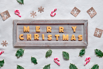 ext Merry Christmas made with cookies on rustic wooden board. Geometric flat lay on white textile background. Zero waste decor from craft paper , candy canes and natural holly leaves.