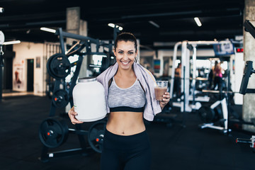 Fototapeta na wymiar Young attractive woman after successful workout in modern fitness gym holding big white protein jar and drinks shake with drinking straw in other hand.