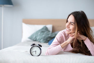 Smiling young woman lying awake in bed with alarm clock, good morning.
