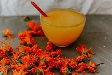 A small cup is filled to the top with yellow liquid honey. Medicinal herbal dried plants marigold, orange calendula. Neutral white and gray background.
