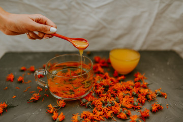 A woman's hand pours honey from a spoon into a cup Flower tea in a large transparent mug.A small...
