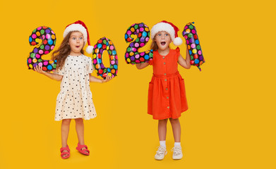 Cheerful children in Santa hats holding colorfull numbers 2021  on yellow background. Happy New Year party with balloons