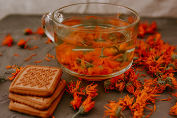 Tea with calendula flowers and biscuits. Transparent glass cup and saucer. Medicinal herbal dried plants marigold, orange calendula. Neutral white and gray background.