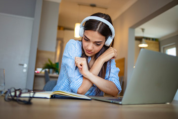 Millennial girl in wireless headphones sit at desk at home working on modern laptop, young ethnic...