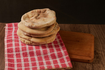 Pita breads or arabian bread stacked on a rustic cloth and a cutting board on a wooden table with black background