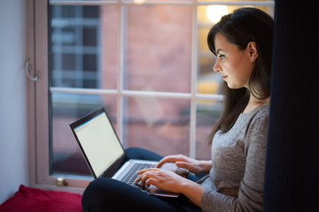 A woman uses her laptop while sitting by the window in the living room of a flat in Edinburgh, Scotland, UK, where lights from other flats can be seen on the background