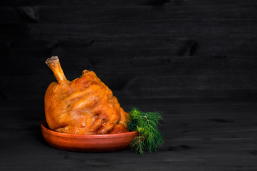Baked pork knuckle with dill greens in a clay pan on a dark wooden background