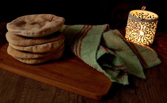 several pita breads or arabian bread stacked on a rustic cloth and a cutting board on a wooden table with black background with soft candle light