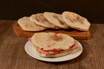 Pita bread sandwiches with ham and tomatoes. the sandwich served on a white earthenware plate and on a wooden board there are more breads.
