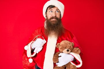 Handsome young red head man with long beard wearing santa claus costume holding teddy bear smiling looking to the side and staring away thinking.