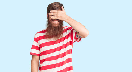 Handsome young red head man with long beard wearing casual clothes smiling and laughing with hand on face covering eyes for surprise. blind concept.