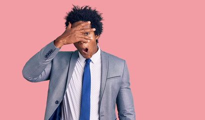 Handsome african american man with afro hair wearing business jacket peeking in shock covering face and eyes with hand, looking through fingers with embarrassed expression.