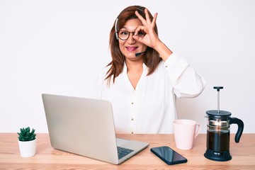 Middle age brunette hispanic woman working at the office wearing operator headset smiling happy doing ok sign with hand on eye looking through fingers