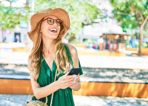 Young beautiful blonde woman on vacation wearing summer hat and glasses smiling happy. Standing with smile on face using smartphone at street of city.