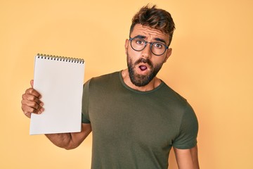 Young hispanic man wearing glasses holding notebook scared and amazed with open mouth for surprise, disbelief face