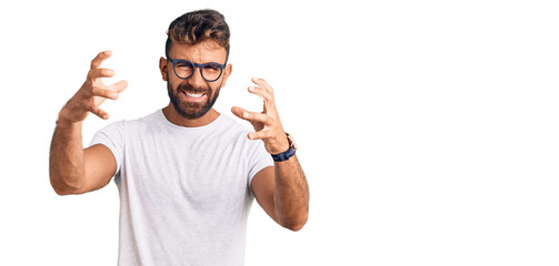 Young hispanic man wearing casual clothes and glasses shouting frustrated with rage, hands trying to strangle, yelling mad