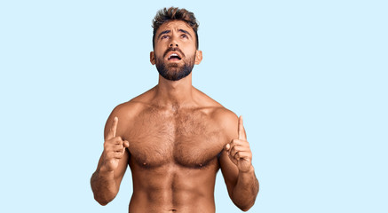 Young hispanic man wearing swimwear shirtless amazed and surprised looking up and pointing with fingers and raised arms.