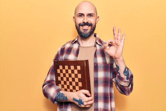 Young handsome man holding chess doing ok sign with fingers, smiling friendly gesturing excellent symbol