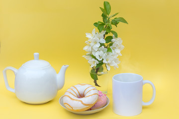 On a yellow background, a white mug with a hot drink, a kettle, a branch of a blossoming apple tree in a vase and donuts. Idea - Teapot and mug without logos - advertising space. Morning coffee