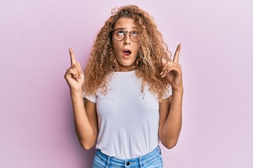 Beautiful caucasian teenager girl wearing white t-shirt over pink background amazed and surprised looking up and pointing with fingers and raised arms.