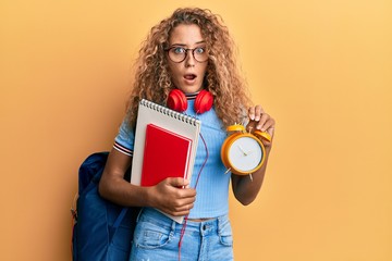 Beautiful caucasian teenager girl wearing student backpack and holding alarm clock in shock face, looking skeptical and sarcastic, surprised with open mouth