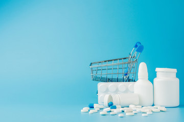scattered variety pills, drugs, spay, bottles, thermometer, syringe and empty shopping trolley cart on blue background. pharmacy shopping concept