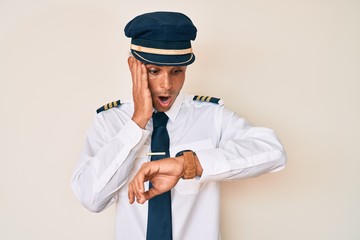 Young hispanic man wearing airplane pilot uniform looking at the watch time worried, afraid of getting late