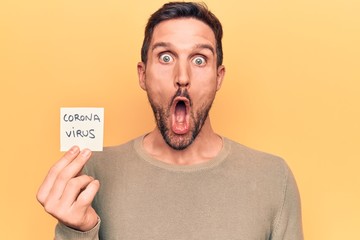 Young handsome man holding reminder with coronavirus message over yellow background scared and amazed with open mouth for surprise, disbelief face