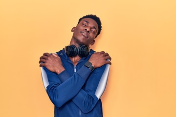 Young african american man wearing sportswear and headphones hugging oneself happy and positive, smiling confident. self love and self care