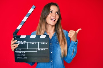 Beautiful caucasian woman holding video film clapboard pointing thumb up to the side smiling happy...